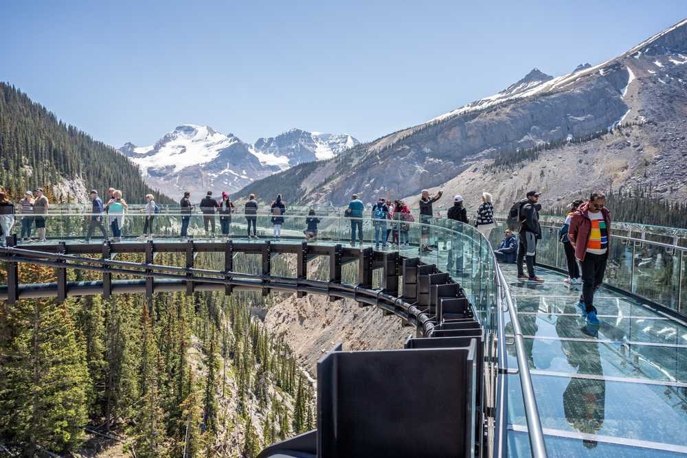 People walking on the Columbia Icefield Skywalk, a glass-enclosed observation bridge overlooking a mountainous landscape with snow-capped peaks and green forests below.