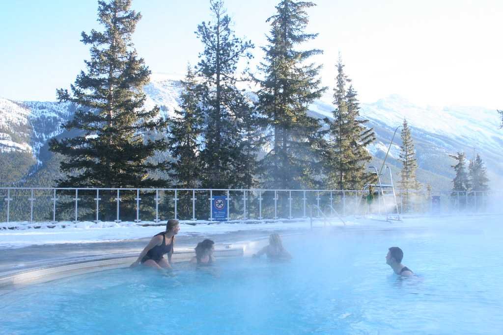 People enjoying a hot spring pool with steam rising, surrounded by tall evergreen trees and snow-covered mountains in the background. This serene setting is one of the many breathtaking Hot Springs in Alberta.