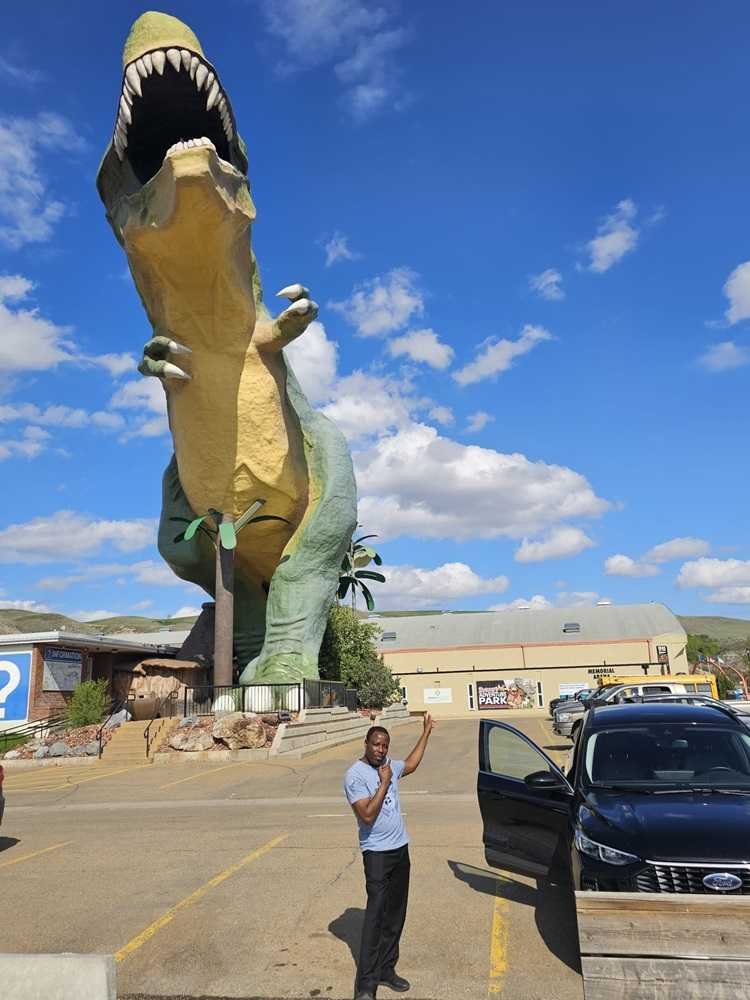 A person stands beside an open car door, posing in front of a large Tyrannosaurus rex statue in a parking lot under a cloudy blue sky, embodying the adventurous spirit we cherish and celebrate in our "About Us" story.