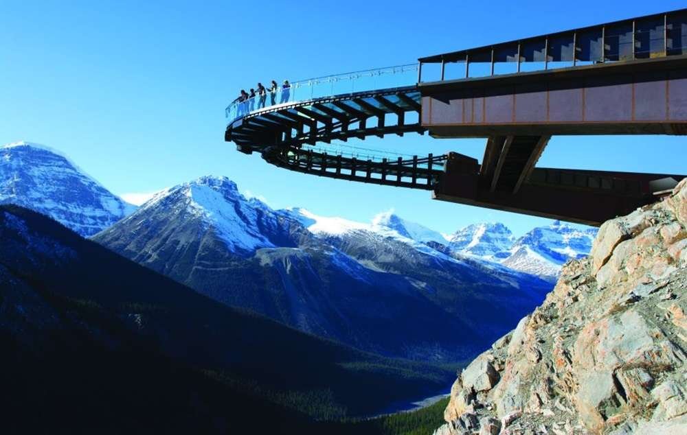 People stand on the glass-floored Columbia Icefield Skywalk, a lookout platform extending from a cliff, offering breathtaking views of snow-capped peaks and the Athabasca Glacier Adventure.