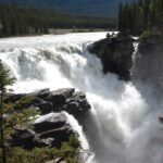 A powerful river waterfall cascades over rocky cliffs surrounded by dense forests and mountains in the background, reminiscent of the breathtaking Athabasca Falls in Jasper National Park.