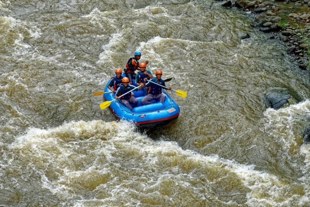 A group of five people wearing helmets and life jackets navigate a blue inflatable raft through rough river rapids with paddles.