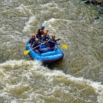 A group of five people wearing helmets and life jackets navigate a blue inflatable raft through rough river rapids with paddles.